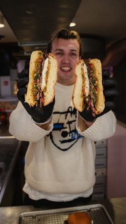 Two homemade smash burgers held by a person wearing black gloves inside a burger restaurant kitchen.