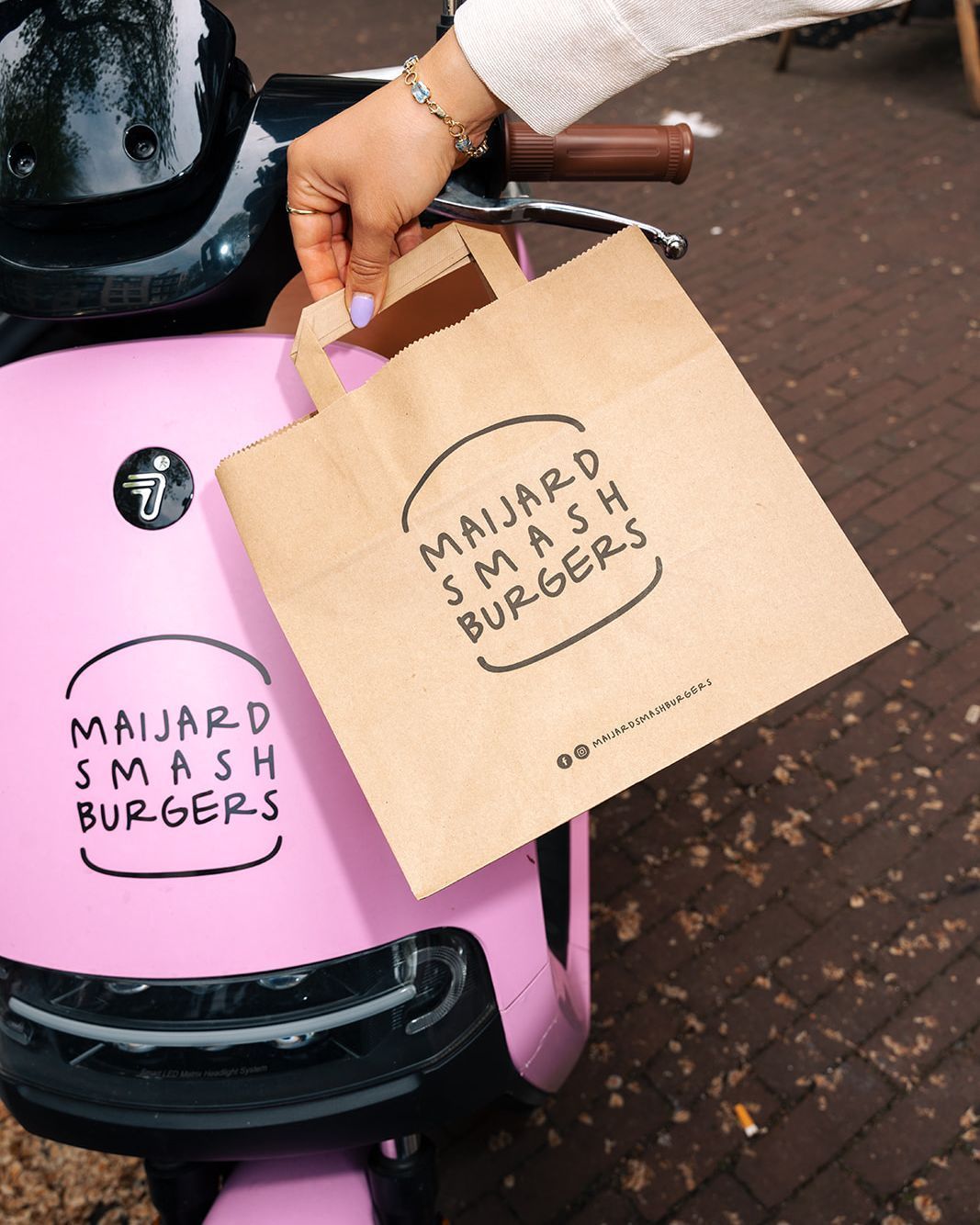 Brown paper bag with Maijard Smashburgers logo held above a pink scooter with same logo on the front panel.