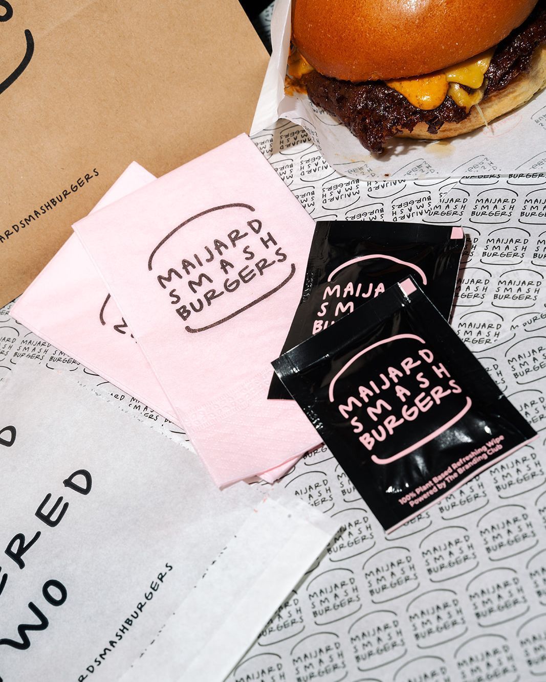 Ketchup packets and pink napkins with Maijard Smashburgers logo beside a cheeseburger on branded paper in Amsterdam.