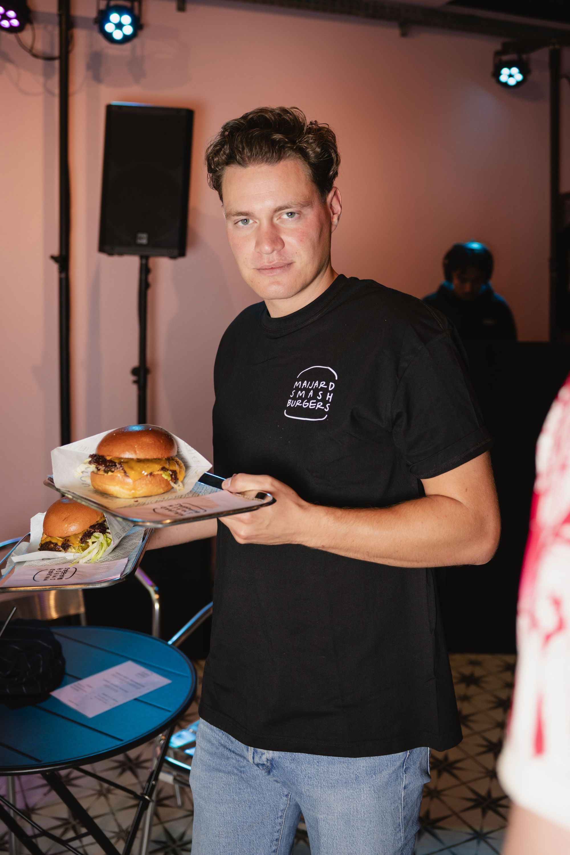 Staff member at Maijard Smashburgers holding plates with homemade smash burgers inside a restaurant setting.