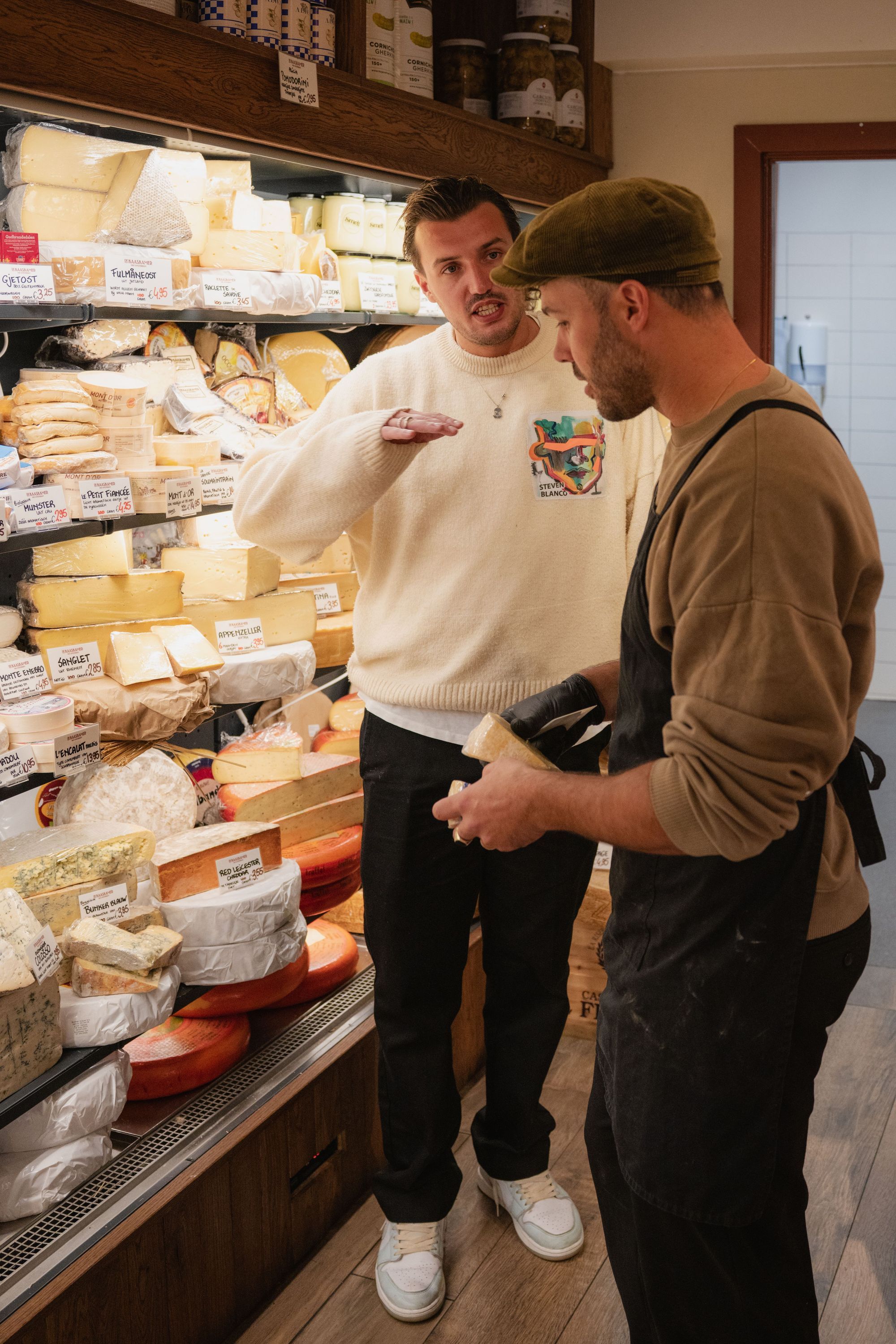 Two men talking near a display of assorted cheeses in a shop, one holding a block of cheese.