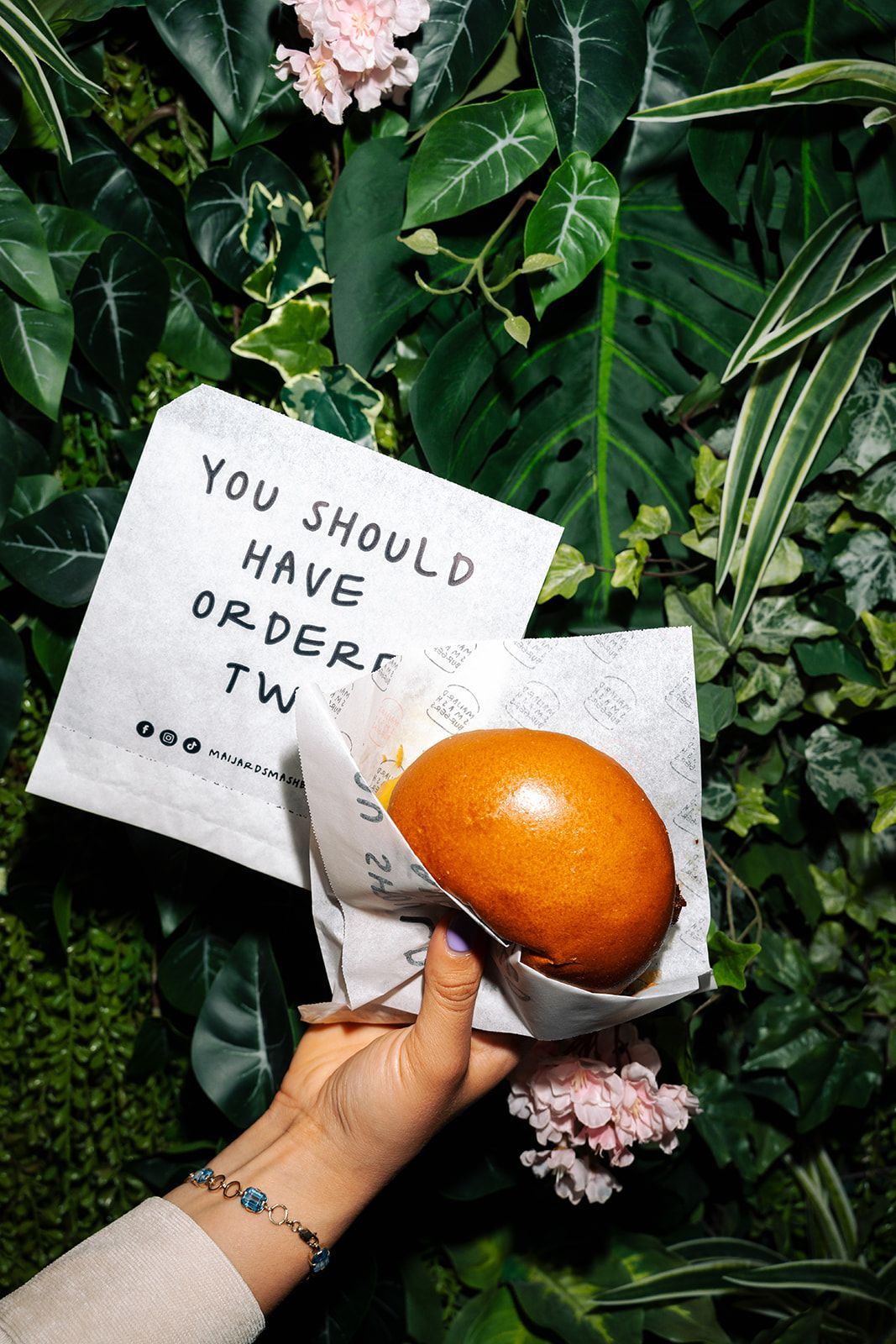Hand holding a wrapped burger with a brown bun in front of a leafy green background and a paper sign with text.