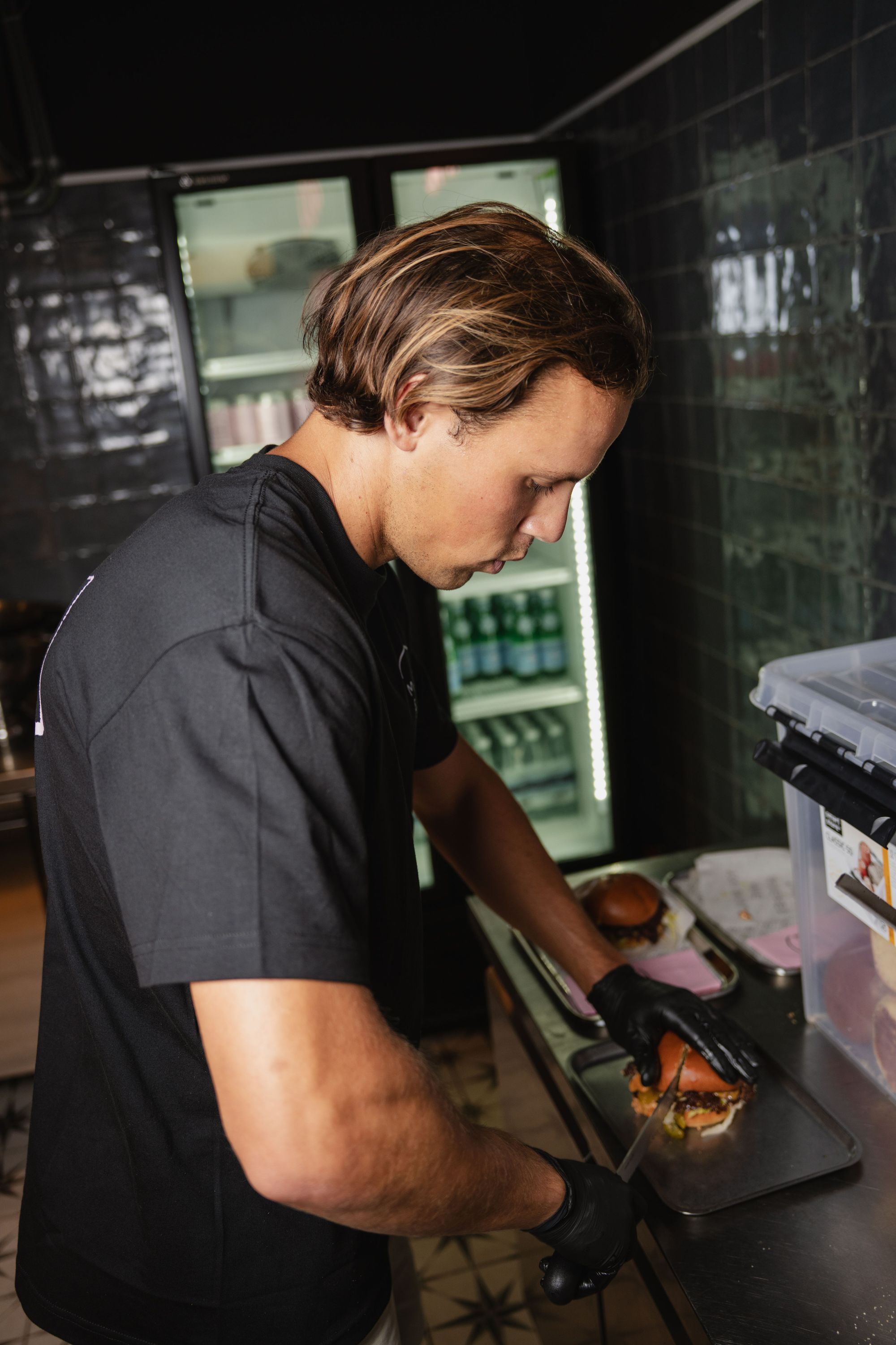 A person wearing black gloves assembling a homemade burger at Maijard Smashburgers kitchen near a refrigerator with drinks.